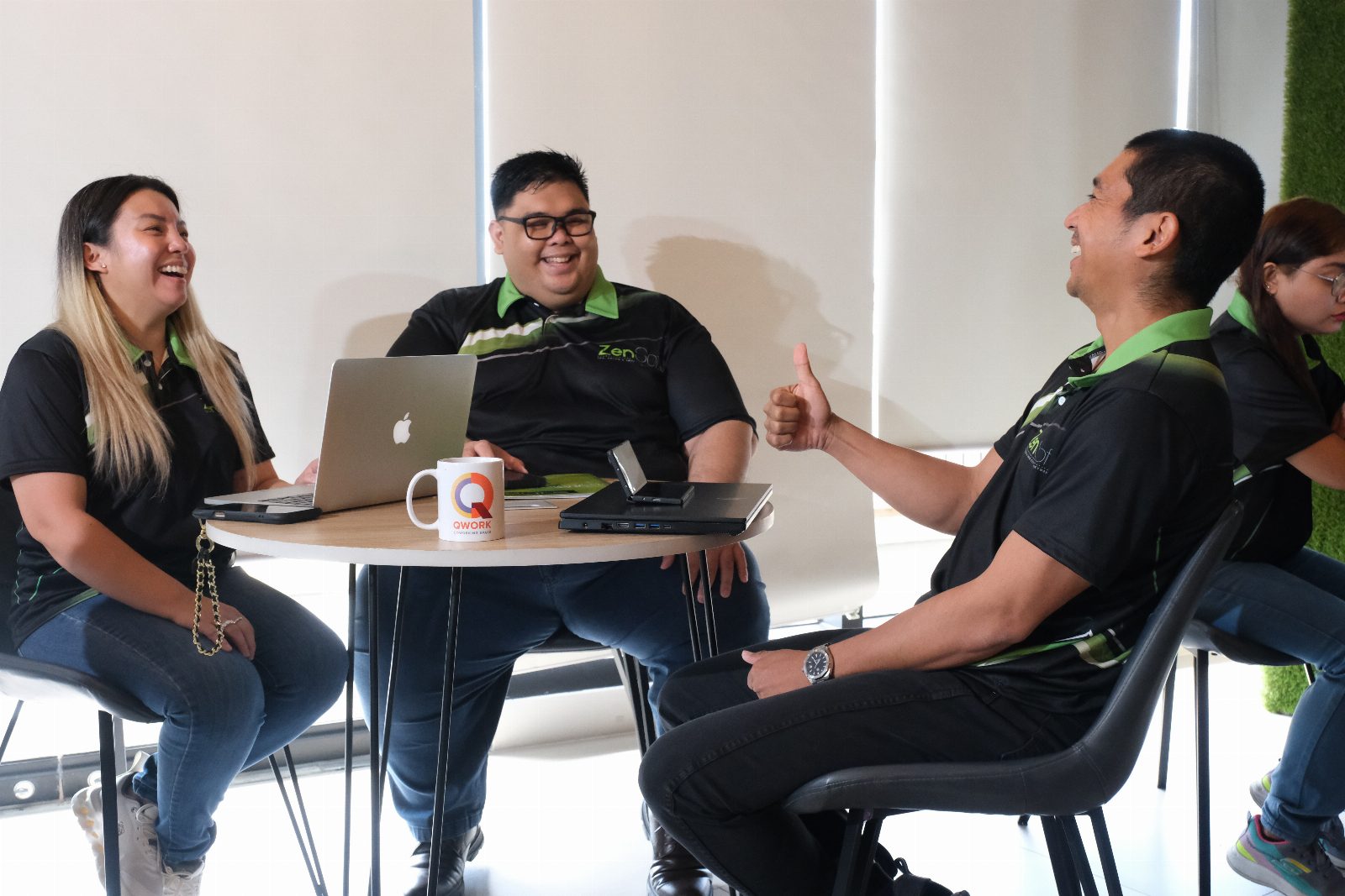 Four ZenSoft team members laughing around a lounge table, one giving a thumbs up, a laptop and coffee mug between them